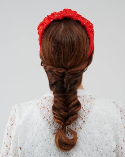 Rear view of a model with auburn hair in a braid, adorned with the Watermelon Headband in red with white polka dots, against the delicate lace backdrop of the Maria Lily Mini-Dress.