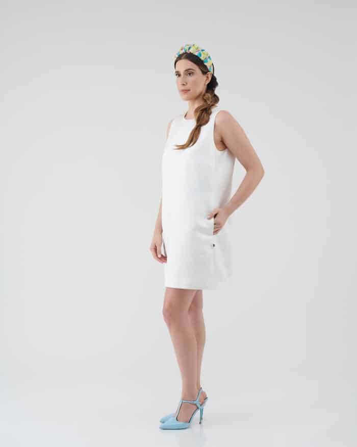 Model poses in a crisp white Maria Daisy Mini Dress and colorful Tulip Headband, paired with chic light blue heels against a clean, white background.