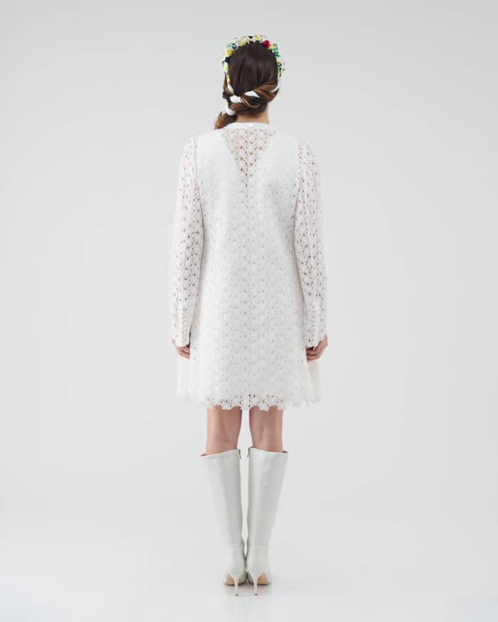 Rear view of a model in a white lace Maria Lily Mini Dress with intricate sequin embroidery and deep V-back, paired with a Lemon Headband and white boots.