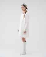 Side profile of a model wearing a white lace Maria Lily Mini Dress with sequin details and silk satin trims, paired with a multicolored Lemon Headband and white knee-high boots.