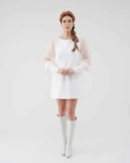 A woman stands facing the camera wearing the Maria Peony Mini Dress, a white dress with sheer balloon sleeves, paired with the vibrant Fern Headband and white knee-high boots