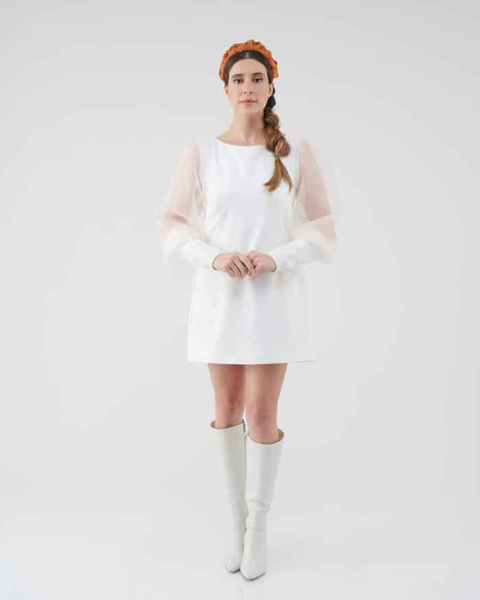 A woman stands facing the camera wearing the Maria Peony Mini Dress, a white dress with sheer balloon sleeves, paired with the vibrant Fern Headband and white knee-high boots