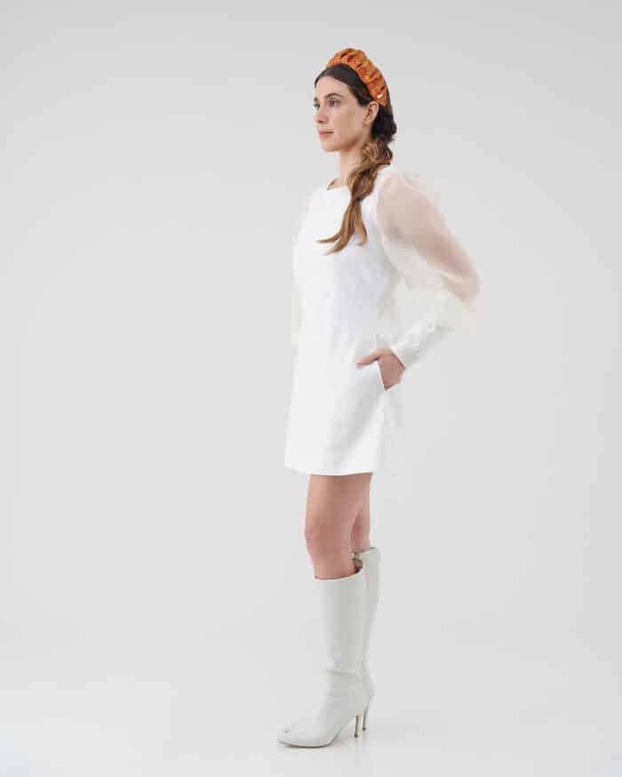 Side profile of a woman modeling the Maria Peony Mini Dress, complete with sheer balloon sleeves and a sleek design, complemented by the Fern Headband and stylish white knee-high boots.