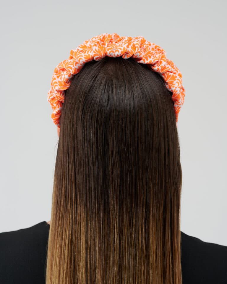 Rear view of a woman wearing the Maria Coral Headband with a rich orange and white floral print, over long, straight brown hair.