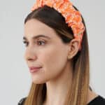 "Side profile of a woman showcasing the Maria Coral Headband with a vibrant orange and white floral pattern on a gathered fabric, complementing her straight long brown hair.