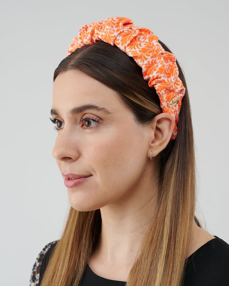 "Side profile of a woman showcasing the Maria Coral Headband with a vibrant orange and white floral pattern on a gathered fabric, complementing her straight long brown hair.