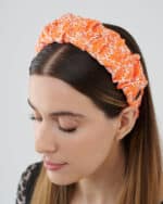 Side view of a woman wearing the Maria Coral Headband, showcasing the vibrant orange and white floral pattern on a fabric headband with a discreet 'Maria' brand tag.
