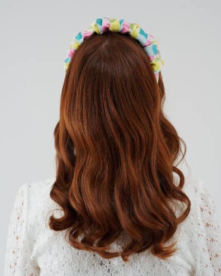 Rear view of a woman with auburn hair wearing the Tulip Headband, featuring a vibrant silk devoré fabric, against the backdrop of the white Maria Lily Mini Dress with lace detail.