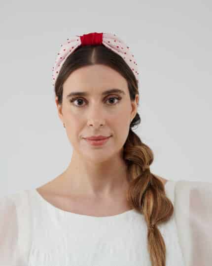 Model facing forward wearing the Strawberry Headband with red crystal bead details and a bold red bow atop a polka-dot fabric, paired with the Maria Jasmine Mini Dress.