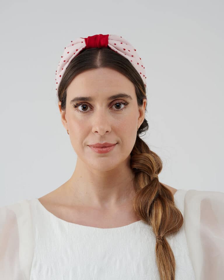 Model facing forward wearing the Strawberry Headband with red crystal bead details and a bold red bow atop a polka-dot fabric, paired with the Maria Jasmine Mini Dress.