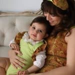 Tender moment of a mother in a brocade dress with orange and lime patterns and Peggy Headband, embracing her baby in a coordinating lime outfit with brocade ruffles.