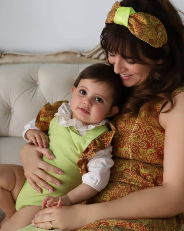 Tender moment of a mother in a brocade dress with orange and lime patterns and Peggy Headband, embracing her baby in a coordinating lime outfit with brocade ruffles.