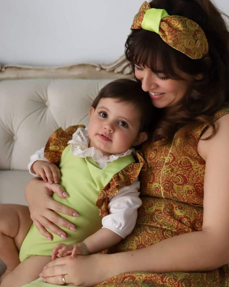 Tender moment of a mother in a brocade dress with orange and lime patterns and Peggy Headband, embracing her baby in a coordinating lime outfit with brocade ruffles.