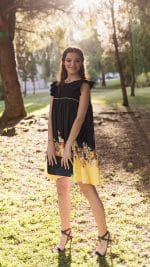 Smiling woman posing in a park at sunset, wearing the Maria Terceira Mini-Dress in black with floral print on yellow hem from Maria Atelier's Summer Special Edition, paired with elegant black heels.