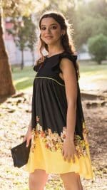Woman modeling the Maria Terceira Mini-Dress in black with floral hem design, from Maria Atelier's Summer Special Edition, holding a clutch and posing outdoors with a sunlit backdrop.