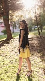 Model turning with a smile, wearing the Maria Terceira Mini-Dress in black with a floral print on the yellow hem, from Maria Atelier's Summer Special Edition, complemented by strappy black heels.