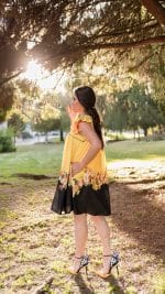 Model posing in a sun-dappled park wearing the Maria Terceira Mini-Dress in yellow with a black floral border from Maria Atelier's Summer Special Edition, paired with black strappy heels.