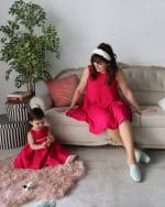 A mother and child in matching fuchsia crepe dresses with white cotton eyelet fabric trims from the Maria Ginger Special Edition Mommy & Me collection, sitting in a stylish living room.