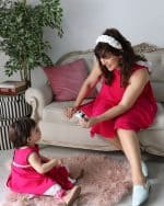 Mother in fuchsia crepe dress and white Daisy Headband smiling at her child, also in a matching fuchsia dress, while playing with a toy in a chic living room.