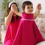 Joyful toddler in a fuchsia dress being playfully lifted by her mother, who is also dressed in a matching fuchsia dress and wearing a white Daisy Headband.