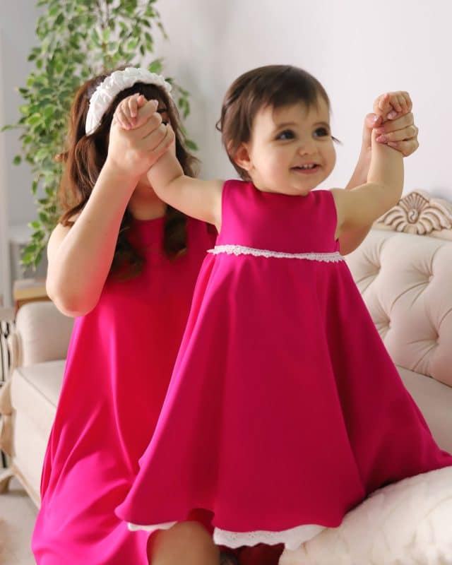 Joyful toddler in a fuchsia dress being playfully lifted by her mother, who is also dressed in a matching fuchsia dress and wearing a white Daisy Headband.