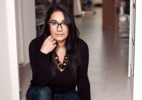 Jaqueline, a confident woman with long dark hair and glasses sitting on the floor, wearing a black V-neck sweater and a chunky necklace, looking directly at the camera with a contemplative expression.