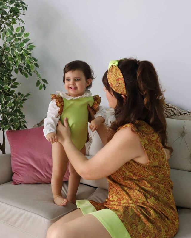 A mother in a brocade dress smiles at her gleeful baby, clad in a matching lime outfit with brocade accents, highlighting their close bond and the intricate design of the Maria Peggy Special Edition Mommy & Me dresses.