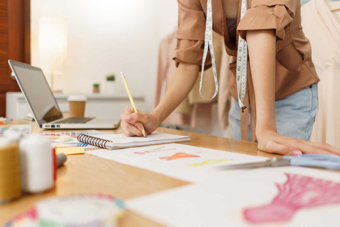 Fashion designer sketching in a notebook on a desk filled with design materials, a laptop open beside her, and a tape measure draped over her shoulders, in a bright workspace.