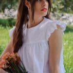 A woman sits under the shade of a tree, wearing a white cotton dress with elegant ruffled shoulders, holding a bouquet of peach flowers. Her gaze is directed slightly upwards, her expression contemplative, complemented by a colorful patterned headband.