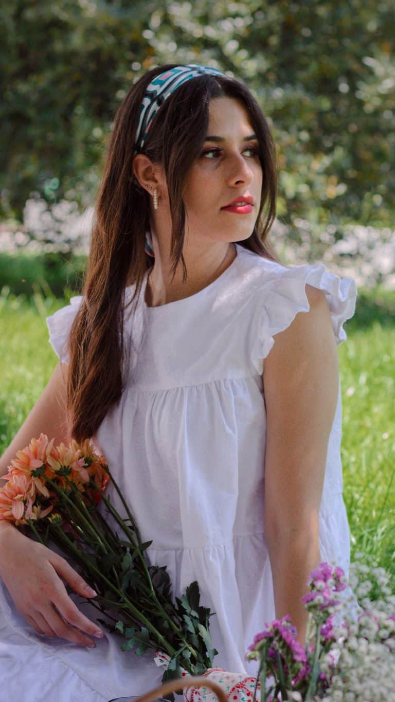A woman sits under the shade of a tree, wearing a white cotton dress with elegant ruffled shoulders, holding a bouquet of peach flowers. Her gaze is directed slightly upwards, her expression contemplative, complemented by a colorful patterned headband.