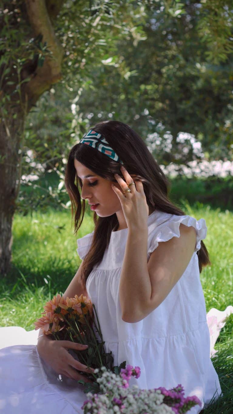 A woman seated outdoors touches her patterned headband and looks down at a bouquet of peach flowers in her lap, wearing a white cotton dress with delicate ruffled shoulders. The dress is detailed with a round neckline and pockets, and the setting is a lush garden with sunlight filtering through the trees.