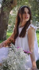A woman in a light, natural white cotton dress adorned with a subtle tone-on-tone star pattern stands outdoors. She gazes off to the side while holding a bouquet of flowers, featuring peach and pink tones. The dress is detailed with a round neckline, pleated ruffle shoulders, and visible layers, enhancing its timeless design.