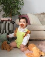 Cute toddler with a pacifier sitting amongst plush toys on a pink rug, wearing a lime green and golden brocade Maria Peggy dress from the Mommy & Me Special Edition, with a decorative indoor plant and elegant sofa in the background.